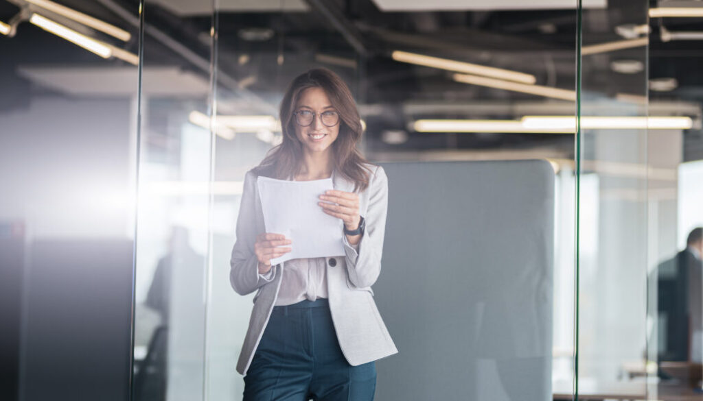 Young smiling businesswoman with documents standing at office and look at camera