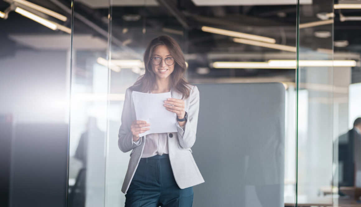 Young smiling businesswoman with documents standing at office and look at camera