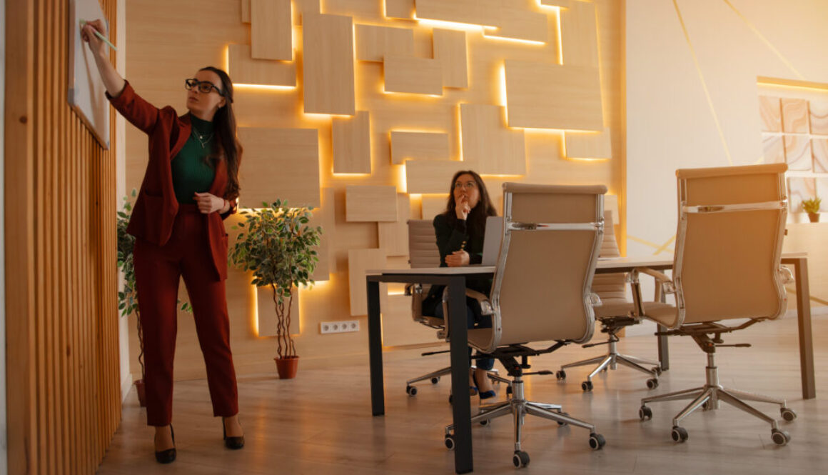 Female professional in red suit writes on whiteboard during a business presentation. Another woman listens attentively at a modern office desk with stylish lighting and decor.